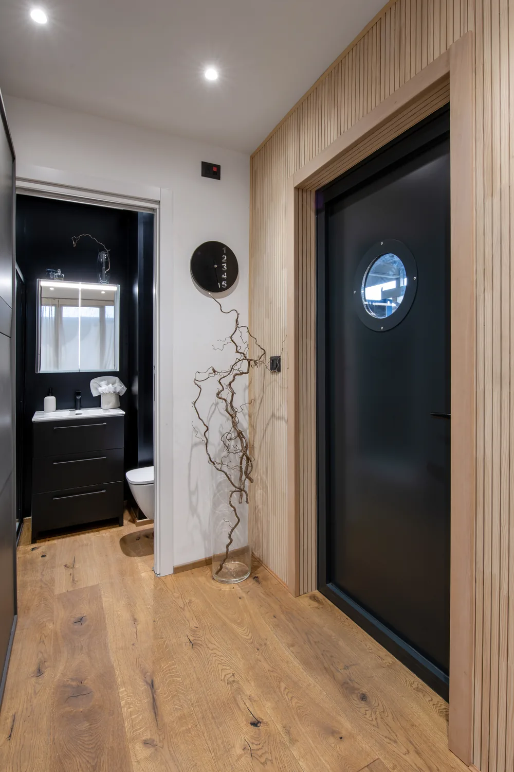 Eingang mit dunkler Tür und Bullauge, Wand aus Holzlatten und links das Bad