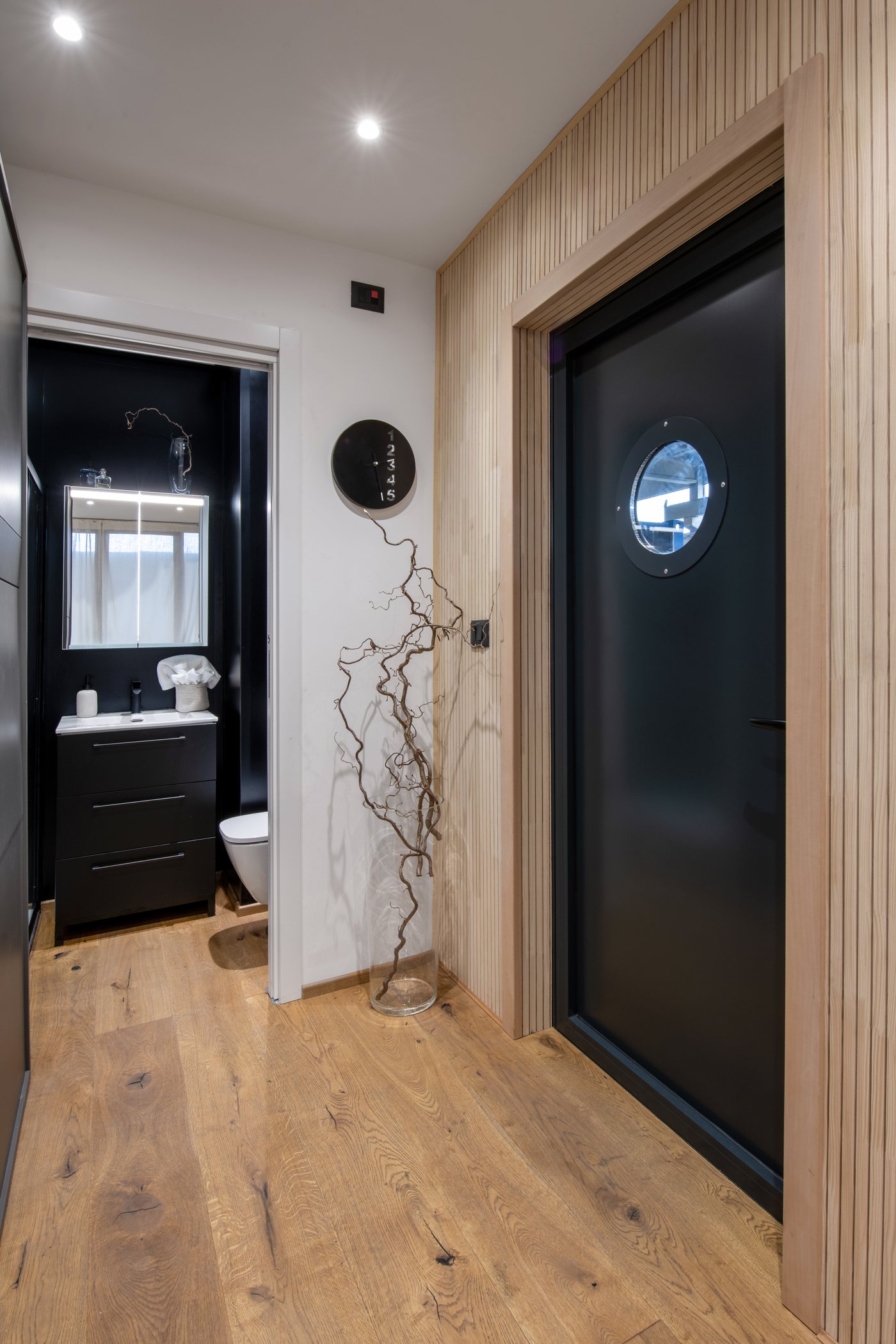 Eingang mit dunkler Tür und Bullauge, Wand aus Holzlatten und links das Bad