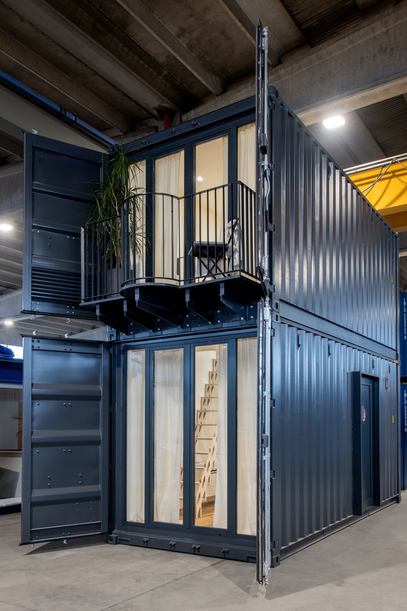 Der zweistöckige Container in Anthrazitgrau mit Balkon und hellen Vorhängen, in der Werkstatt
